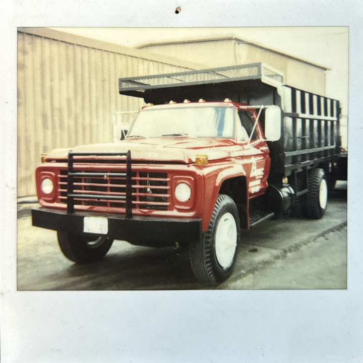 A vintage red truck with a flatbed and a black grille with horizontal bars is parked in front of a beige industrial building. The truck appears well-maintained, and the image has a nostalgic, slightly faded quality typical of older photographs.