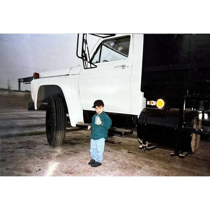 A young child wearing a cap and a blue jacket stands in front of a large white semi-truck. The child is holding a bottle and looks toward the camera, while the truck is parked on a paved area with a road and trees visible in the background.