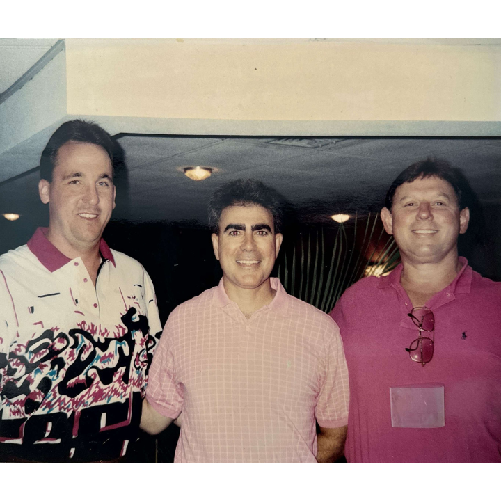 Three men stand together indoors smiling. The man on the left wears a white shirt with abstract patterns, the man in the middle wears a pink polo shirt, and the man on the right wears a red polo shirt with sunglasses hanging from the collar.
