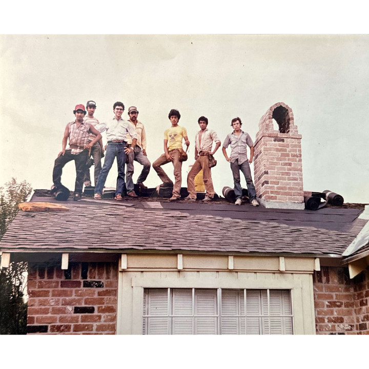A group of eight people stand on the roof of a house next to a brick chimney, posing for the camera. They appear to be construction workers wearing casual clothes and work boots. The sky is clear, and the house features brick and siding exterior with a window below.