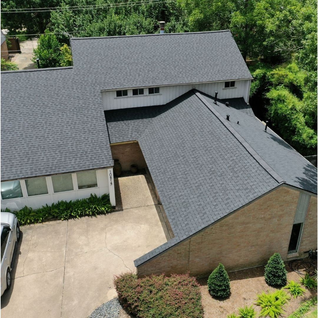 Aerial view of a modern residential house with a dark gray shingle roof, white siding, and beige brick exterior. The driveway is concrete, and the front yard has neatly trimmed bushes and shrubs. The area is surrounded by green trees and foliage.