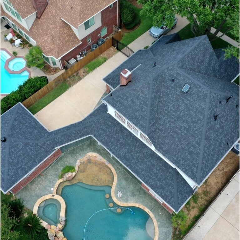 Aerial view of a residential property featuring a house with gray roofing and a backyard pool area. The pool has a curved design with natural stone accents. The adjacent yard is fenced, and neighboring houses with pools are visible in the background.