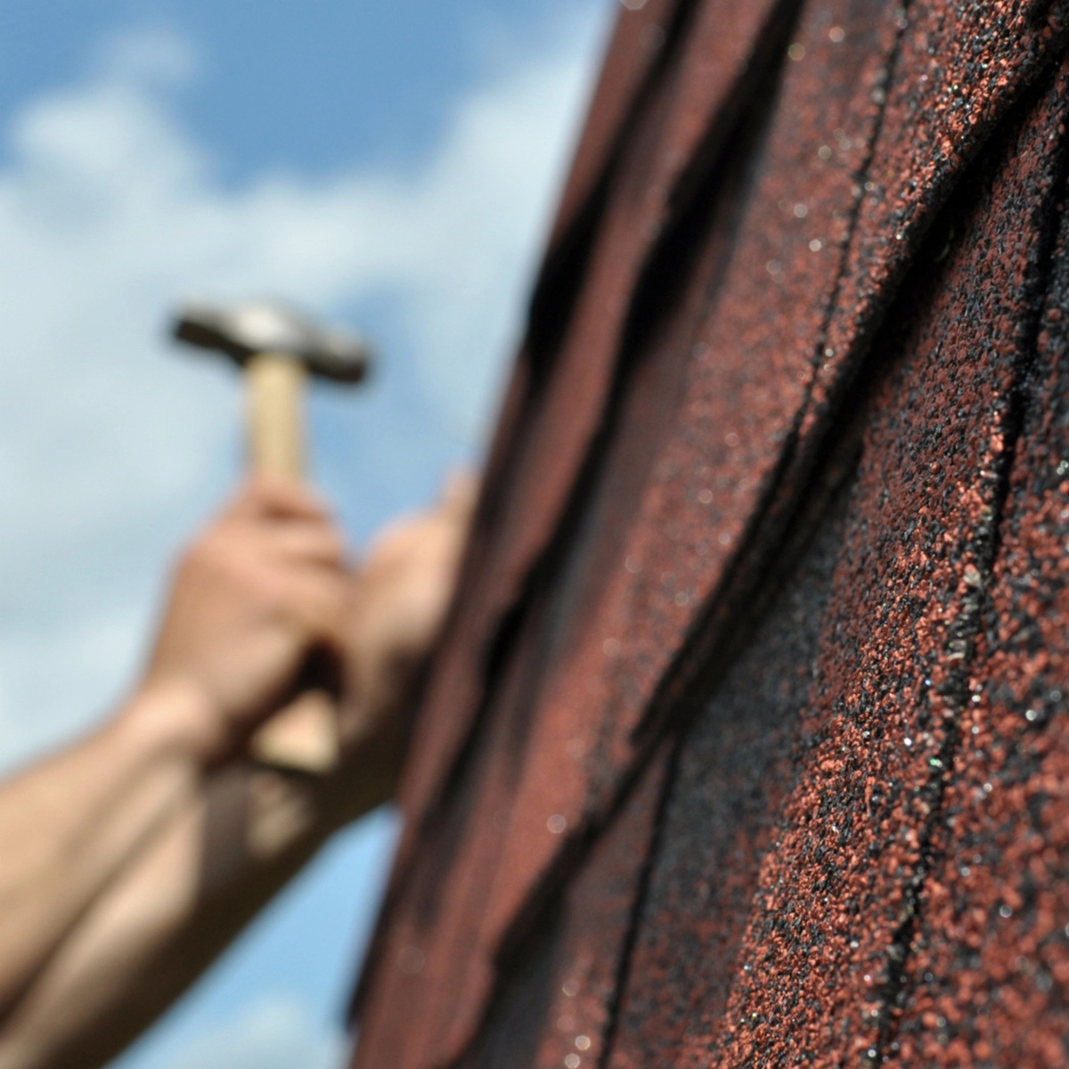 A close-up image shows red roof shingles on the right, with a slightly blurred background of a person using a hammer on the shingles. The sky is blue with some clouds.