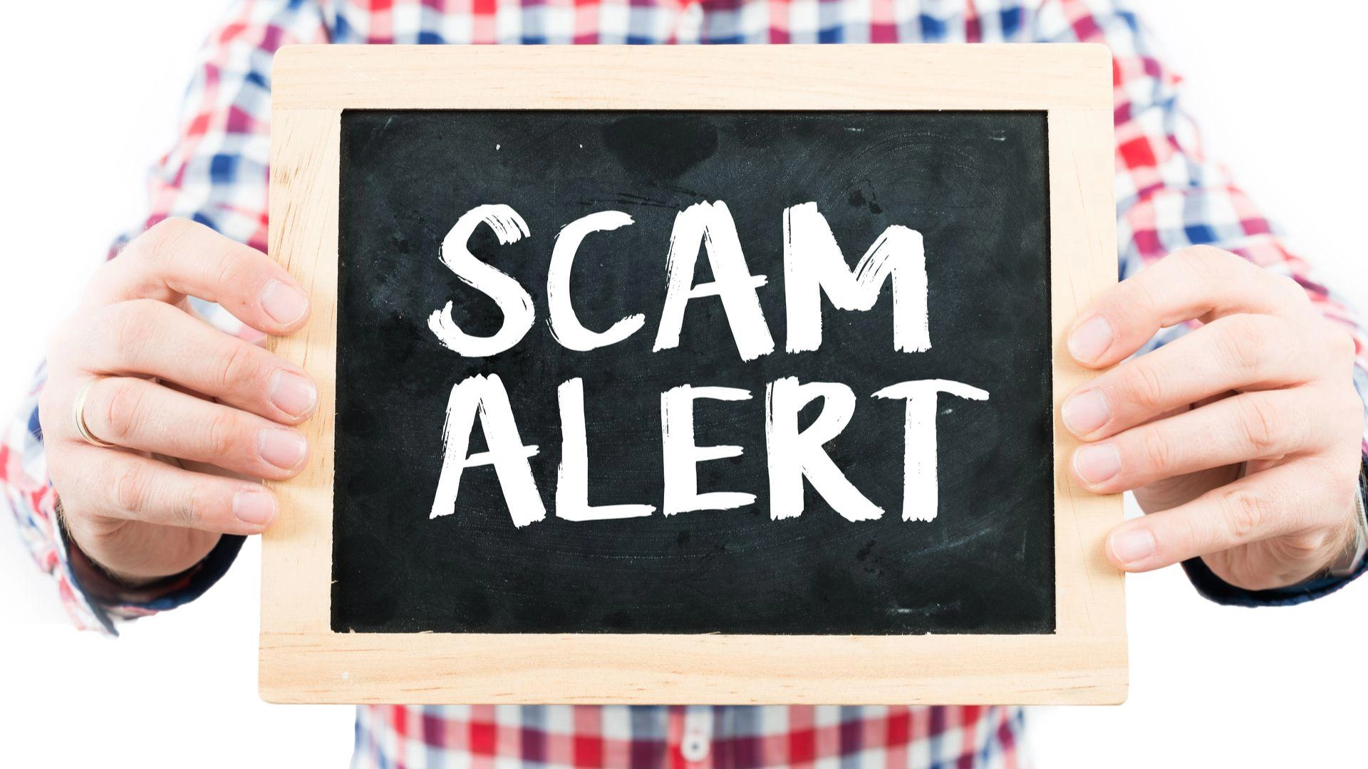 A person wearing a red, white, and blue plaid shirt holds a small blackboard with a wooden frame. The blackboard has "SCAM ALERT" written on it in bold white letters. In the background, you can sense the urgency as this warning could involve anything from online fraud to issues with a Houston roofing company.