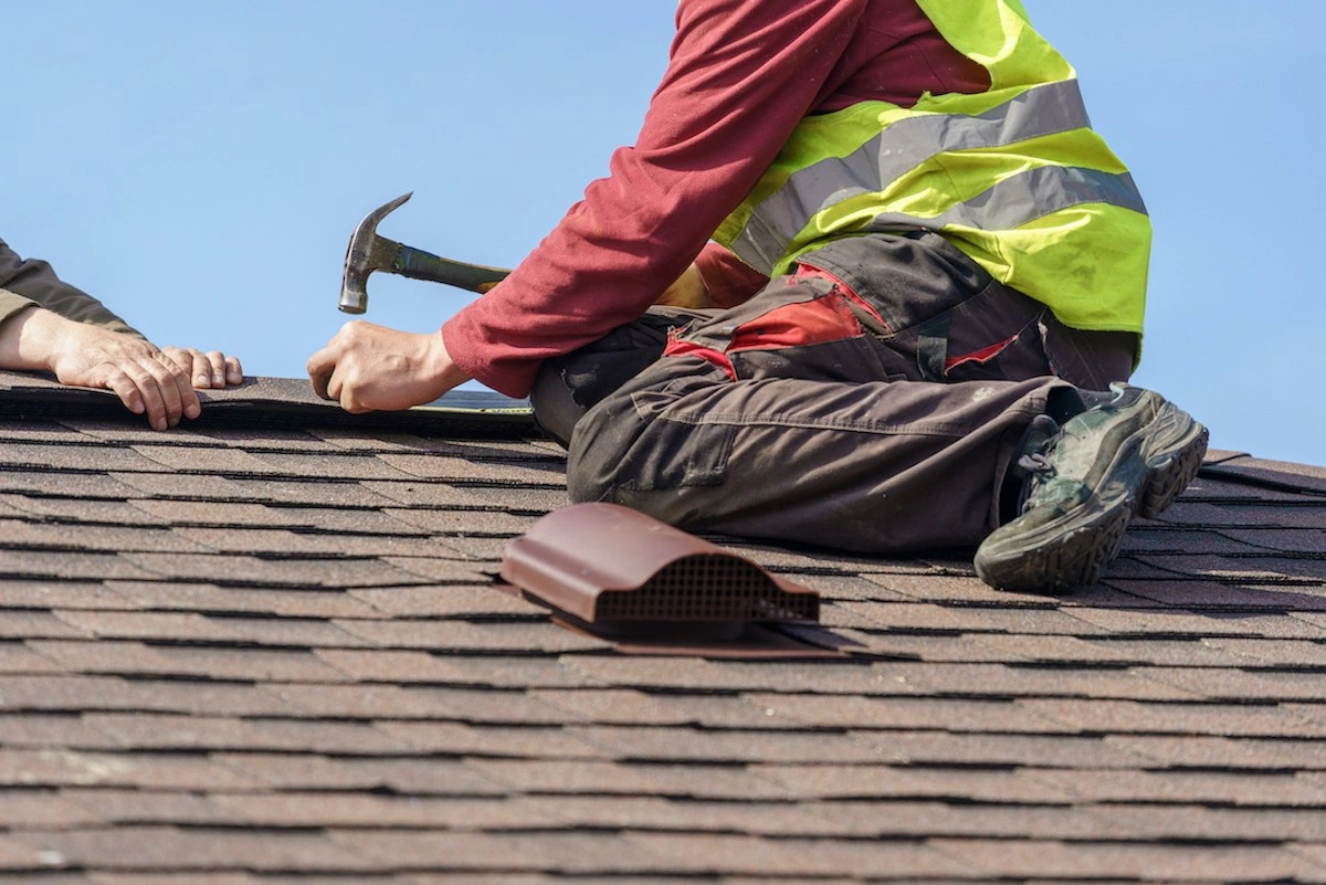 A construction worker in a yellow safety vest and red shirt is kneeling on a shingled roof in Houston, hammering a nail. Another person's hand is assisting by holding a roofing material, while a roof vent is placed nearby.