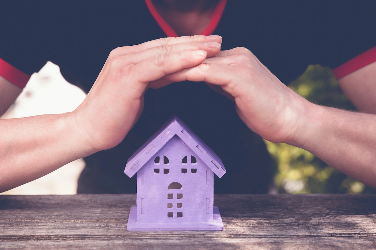 A person in a dark shirt with red trim cups their hands protectively over a small, purple wooden house model on a wooden table. The background, reminiscent of lush Houston greenery, adds depth to the scene.