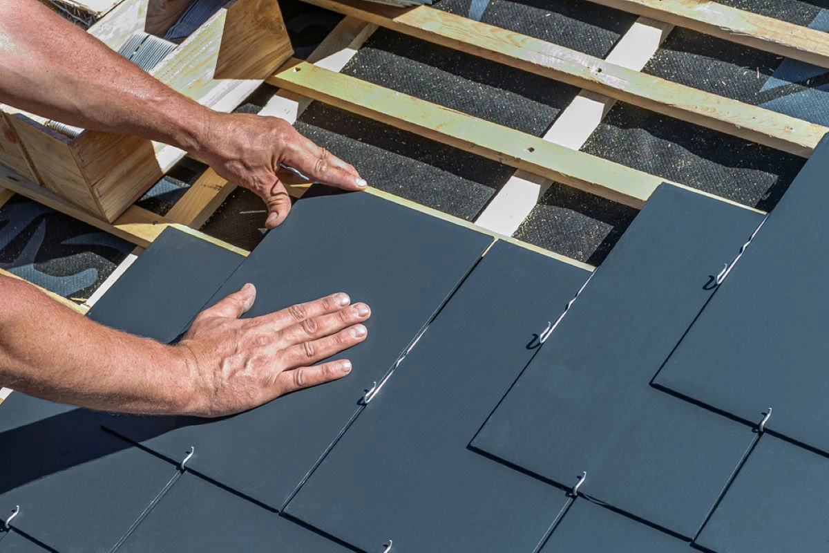 Close-up of a worker's hands installing slate roof tiles on a wooden frame. The tiles are aligned in neat rows, and the worker is positioning a tile, ensuring it fits securely. The background shows other construction materials and partially completed roofing by DaVinci Roofing.