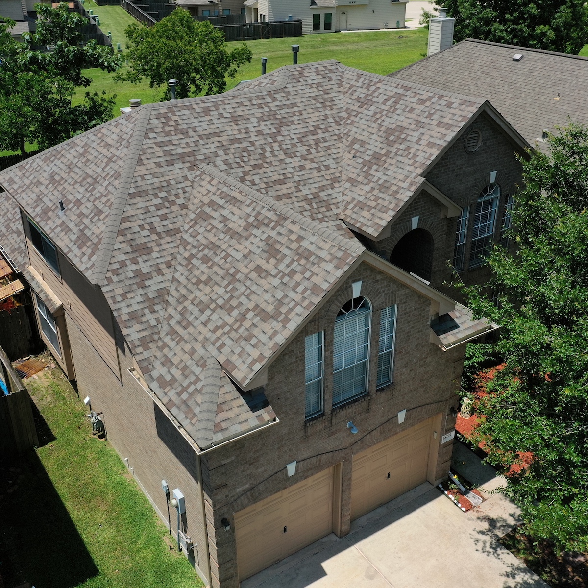 Aerial view of a large two-story brick house in The Woodlands, showcasing a complex, multi-faceted roof covered in gray shingles. The house features an arched window above a double garage and a well-maintained yard with green trees and grass, all benefiting from recent roof replacement efforts.