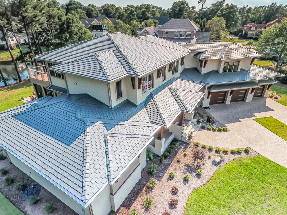 Aerial view of a large, modern two-story house with a blue-tiled roof, multiple gables, and an attached garage. The property features a well-maintained yard with a variety of plants and a driveway leading to the garage. Trees and other homes are visible in the background.