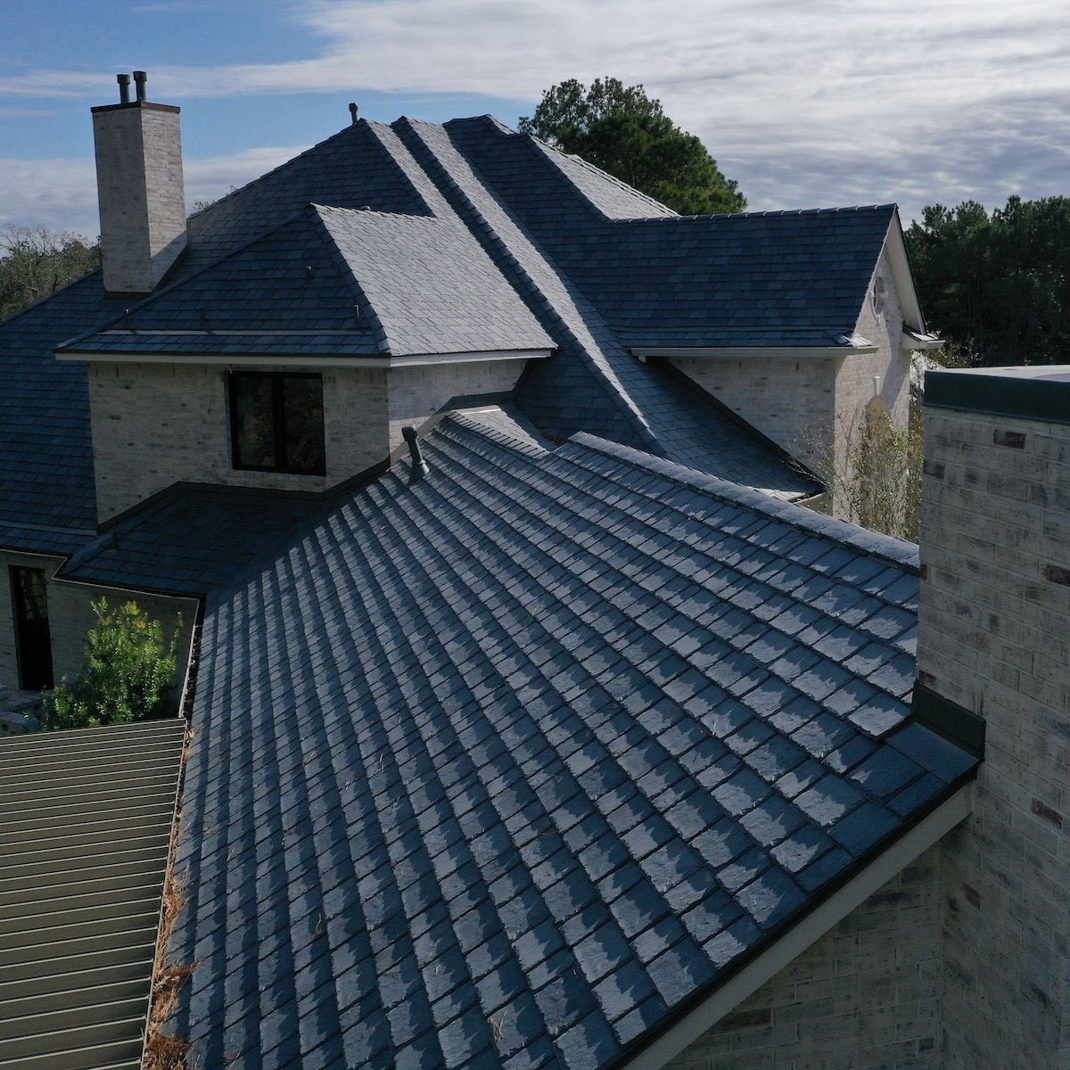 Aerial view of a house in Brays Oaks with a complex roof design featuring grey shingles. The brick exterior walls complement the roofing. The sky above is partly cloudy, creating a mix of sunlight and shadow on the roof and surrounding trees, highlighting the recent roof replacement.