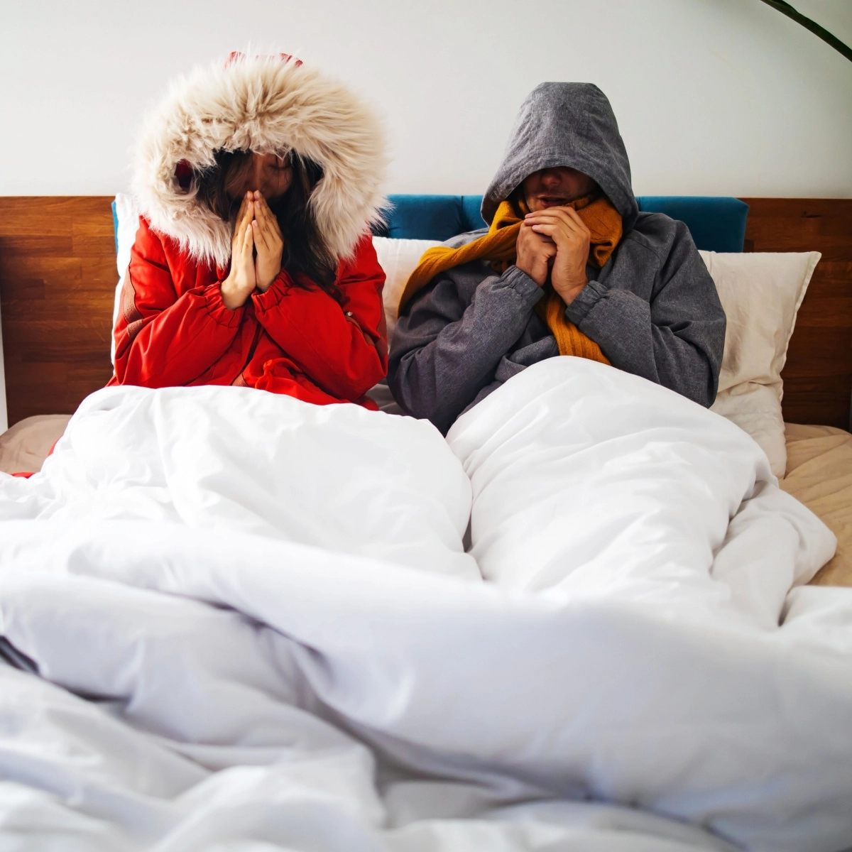 Two people sit in bed under a white comforter, wearing winter clothing. One person wears a red parka with a fur-lined hood, while the other sports a grey hoodie with an orange scarf. Both have their hands close to their faces, appearing to keep warm as if they were escaping from Houston's unexpected roof damage woes.