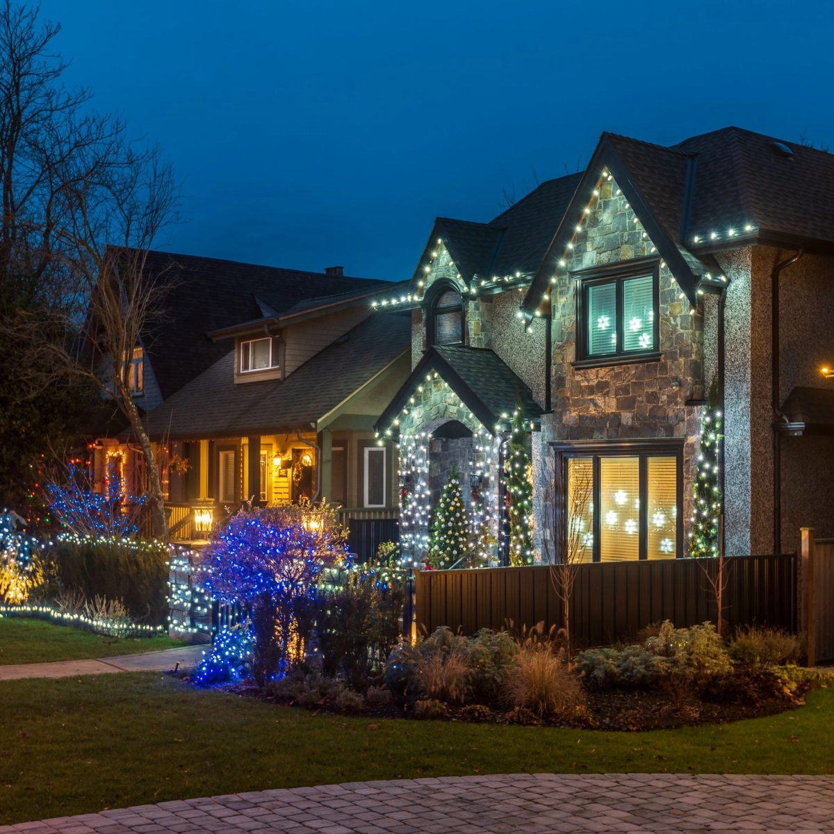 A house is decorated with festive holiday lights at dusk. The lights in various colors outline the roof, windows, and shrubbery, creating a cheerful holiday atmosphere. Thanks to Katy roofing expertise, even the neighboring house is partially visible, similarly adorned with lights.
