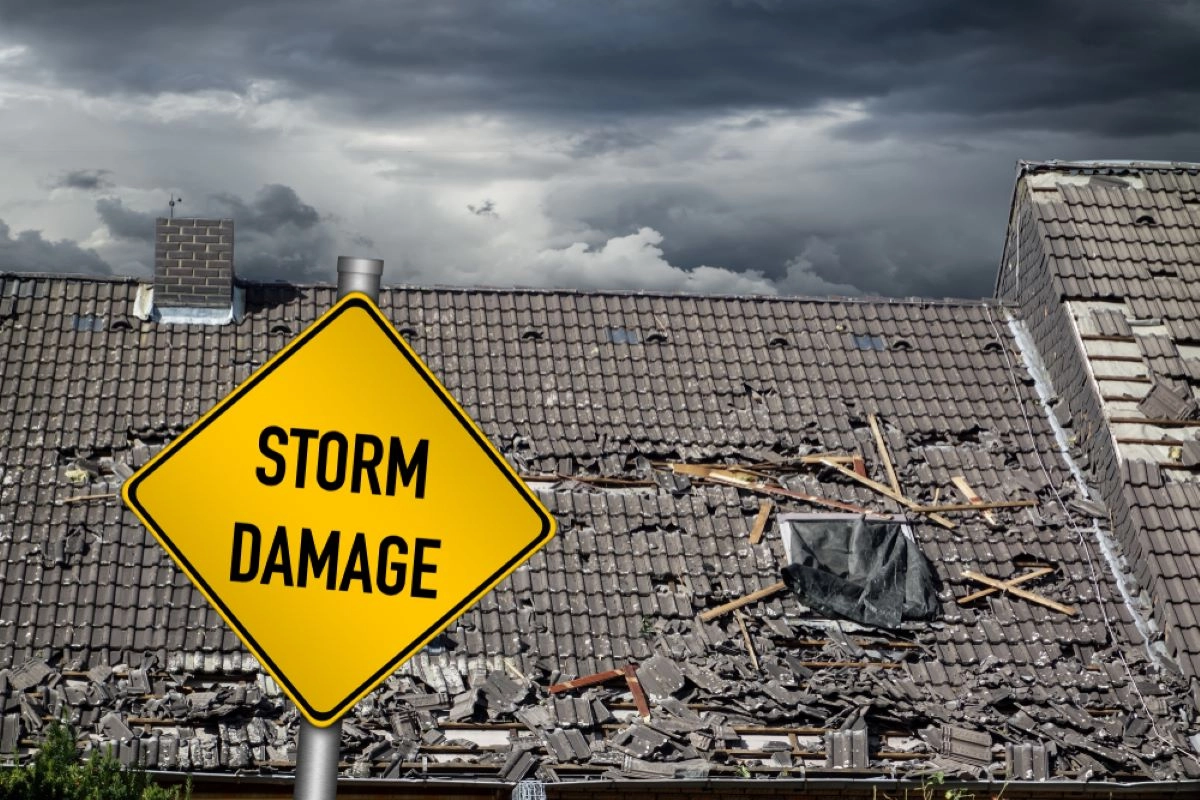 A yellow warning sign reading "STORM DAMAGE" stands in front of a building with a severely damaged roof. The roof's tiles are broken and scattered, indicating Houston hail damage, and the sky above is filled with dark, ominous clouds, suggesting severe weather conditions.