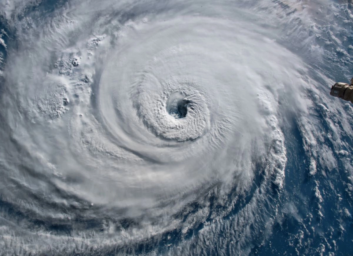 Satellite image of a powerful hurricane swirling over the ocean, with a clearly defined eye in the center. White clouds spiral outward, showing the storm's extensive reach. The blue ocean contrasts with the white, whirling clouds, hinting at potential roof damage as it approaches Houston.