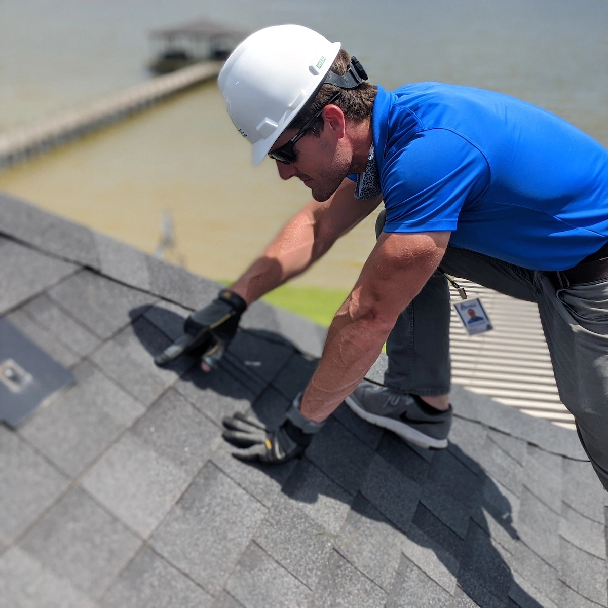 A construction worker in a blue shirt, gray pants, gloves, and a white hard hat is inspecting the shingles on a roof. In the background, there is a body of water and a pier. Leaning forward as part of the meticulous Houston roof repair process, the worker closely examines the surface.