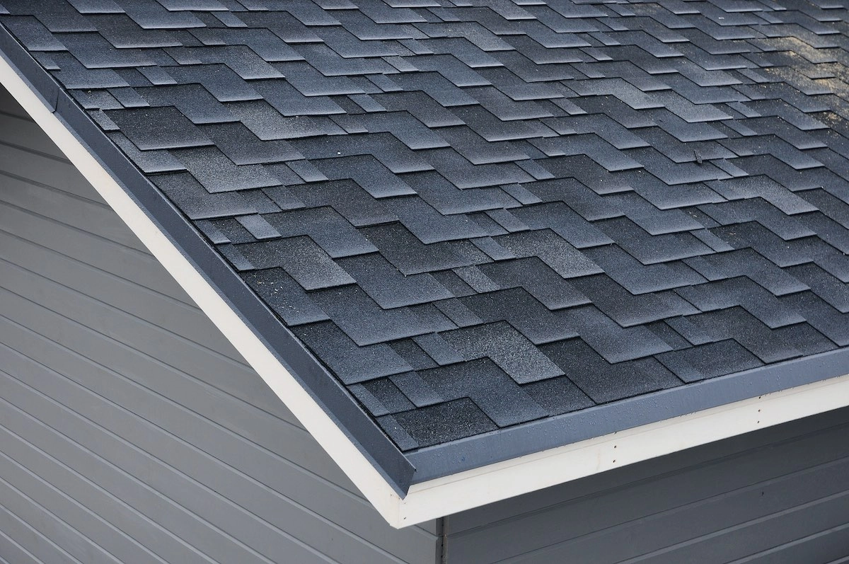 A close-up view of a sloped roof with dark gray asphalt shingles arranged in a zigzag pattern. The roof edge, expertly done by Bellaire Roof Repair, is defined by a white trim, and the siding of the building is visible in shades of gray.