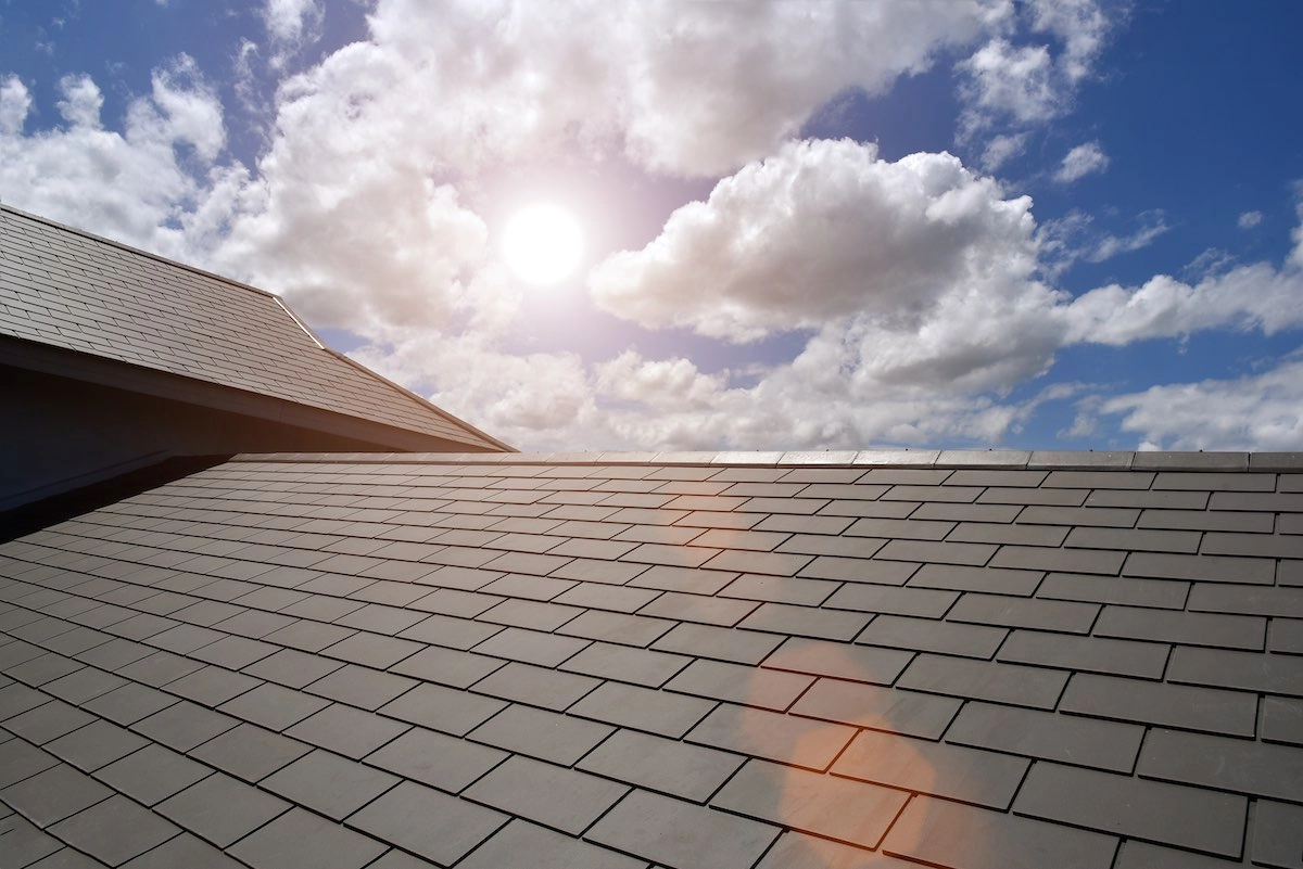 A view of a sloped roof with grey shingles under a bright, sunny sky. White clouds are scattered across the blue sky, and the sun is shining brightly, casting a lens flare over the roof—precisely the kind of quality you'd expect from Houston roof experts.