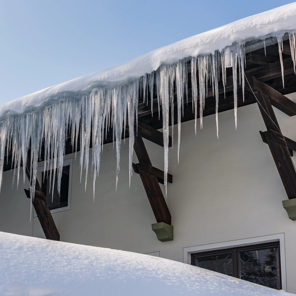 A house with a snow-covered roof, large icicles hanging from the eaves, and the ground piled high with snow showcases exceptional craftsmanship. The building features wooden supports and a couple of windows, standing proudly under a clear blue sky—a scene that would make any Houston roofing expert proud.
