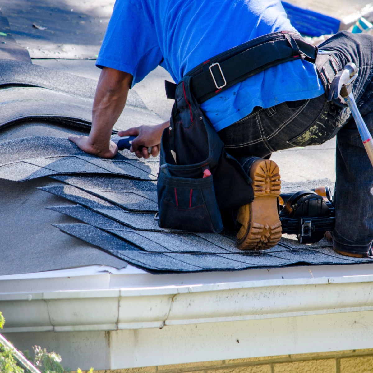 A person in a blue shirt and tool belt is roofing a house in Friendswood. Kneeling on the gray shingled roof, they work with a hammer and tools to install new shingles. The scene includes the gutter and part of the house's exterior wall, illustrating a precise and dedicated Friendswood roof replacement.