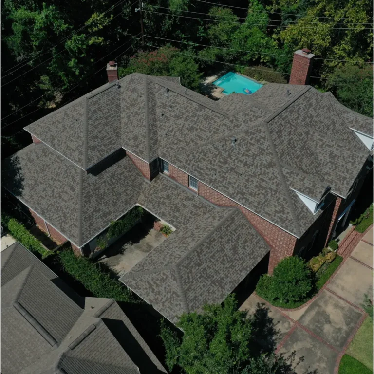 Aerial view of a large two-story house with a complex roof structure, red brick walls, and multiple chimneys. The house is surrounded by lush green trees and shrubbery, with a backyard swimming pool visible in the upper part of the image. Discover top-notch craftsmanship from the best roofing company near you in Houston.