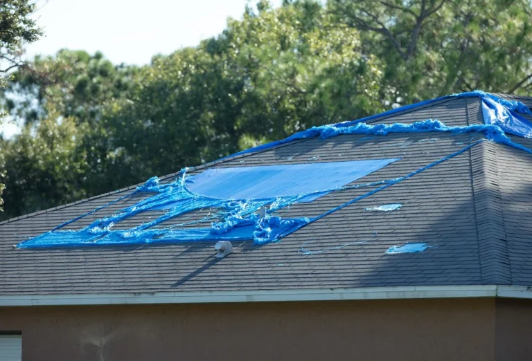 A house with a damaged roof covered by a torn blue tarp. The tarp is partially secured, with sections flapping in the wind. Trees are visible in the background, indicating the house is in a wooded area. The roof appears to have missing or damaged shingles, suggesting it might need a Houston roof replacement soon.