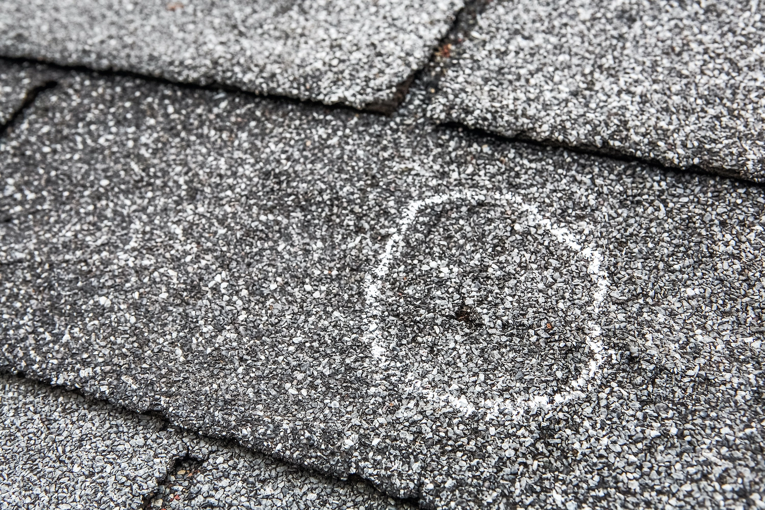 Close-up of a damaged roof shingle marked with white chalk. The dark asphalt shingle has granules for water resistance, but an indentation or hole has been circled to indicate damage needing repair, highlighting the need for professional Houston roof repair services.