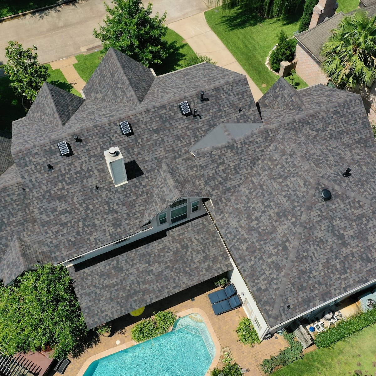 A bird's-eye view of a large house with a dark grey, multi-gabled roof in Meadows Place. The backyard features a swimming pool surrounded by a patio with lush greenery and adjacent trees. Several vents and a chimney, likely part of the recent roof replacement, are visible on the roof.