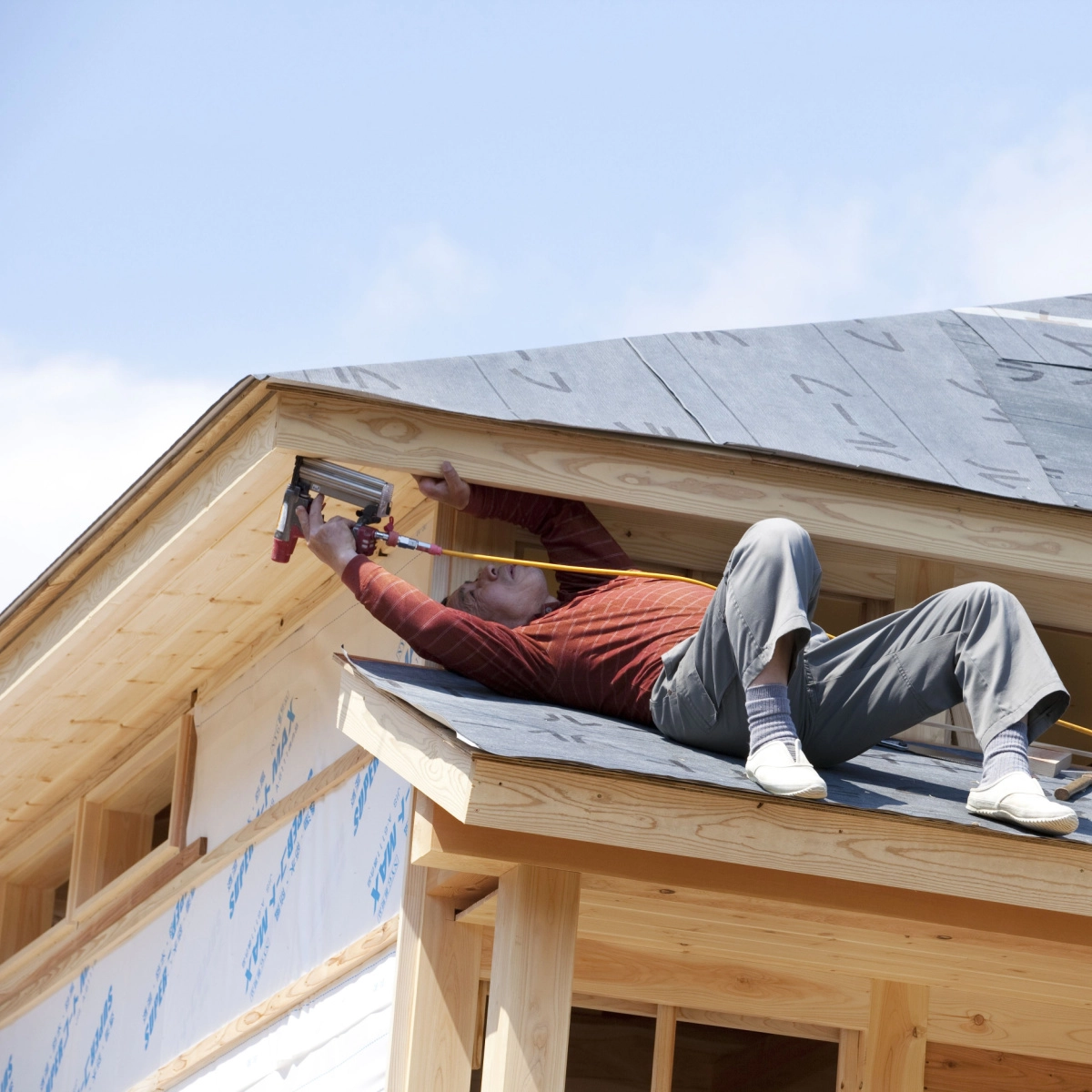 A person wearing a red shirt and gray pants is lying on the roof of a house under construction. They are using a staple gun to secure a piece of wood to the fascia board while holding a measuring tape. The sky is clear with some clouds in the background.