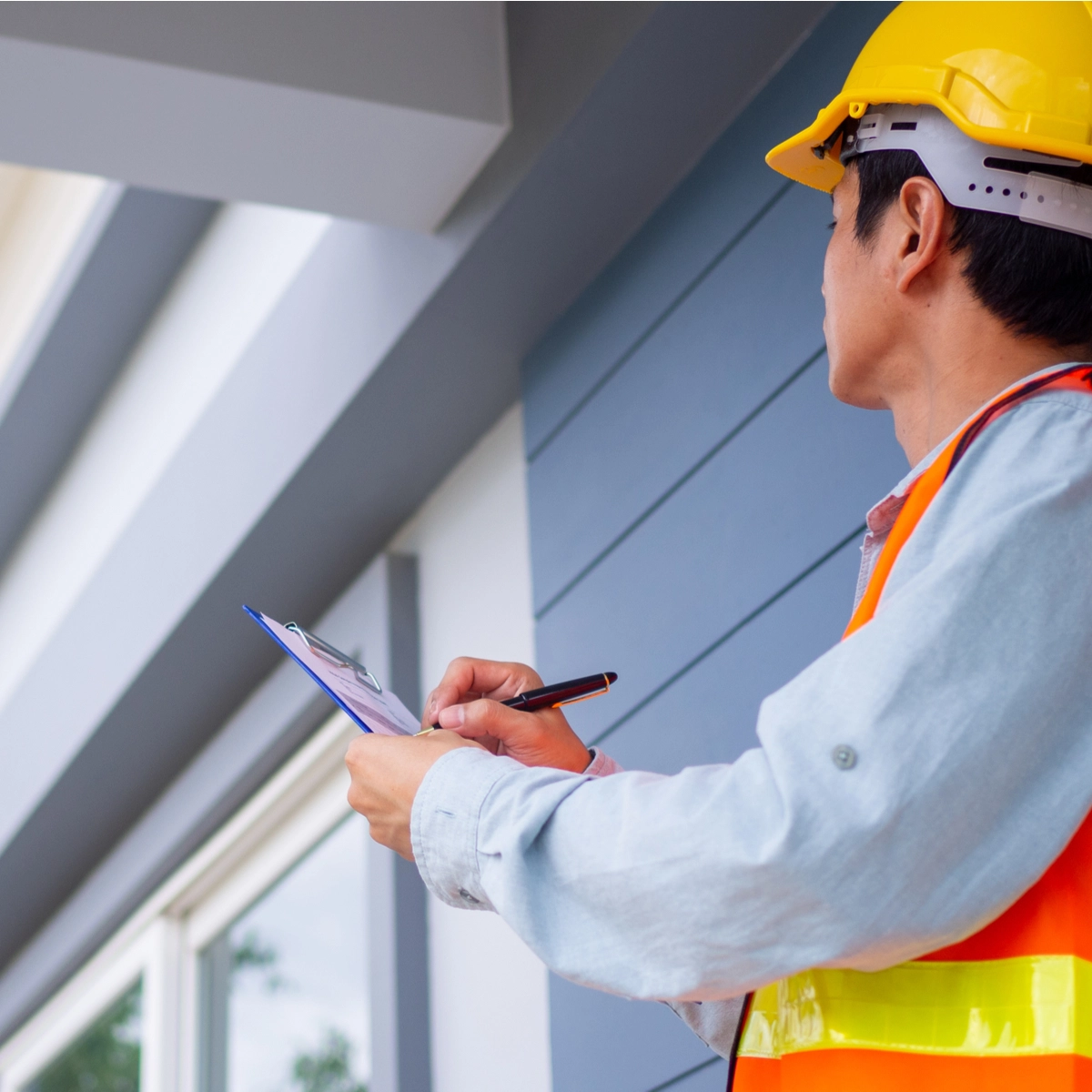 A construction worker in a yellow hard hat and orange safety vest stands outdoors in Pearland, writing on a clipboard. Facing a building with blue siding and the top of a window visible, he assesses potential roof damage following the hurricane.