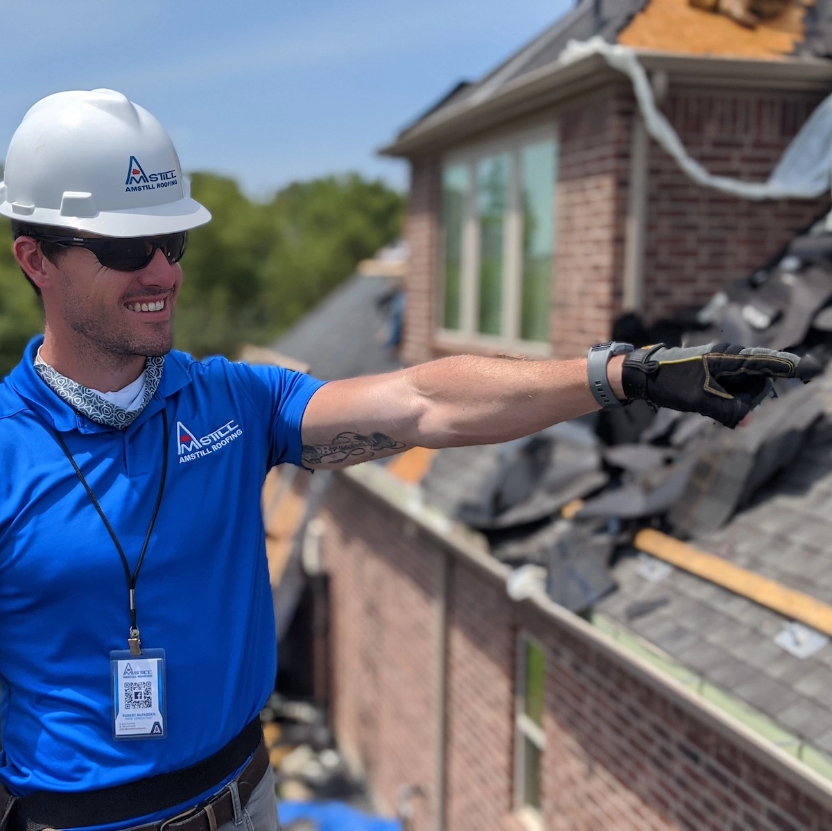 A person wearing a white hard hat, blue polo shirt, and gloves points at something off-camera while standing on the Copperfield Place rooftop. The background shows a building with a partially dismantled roof, indicating an ongoing roof replacement.