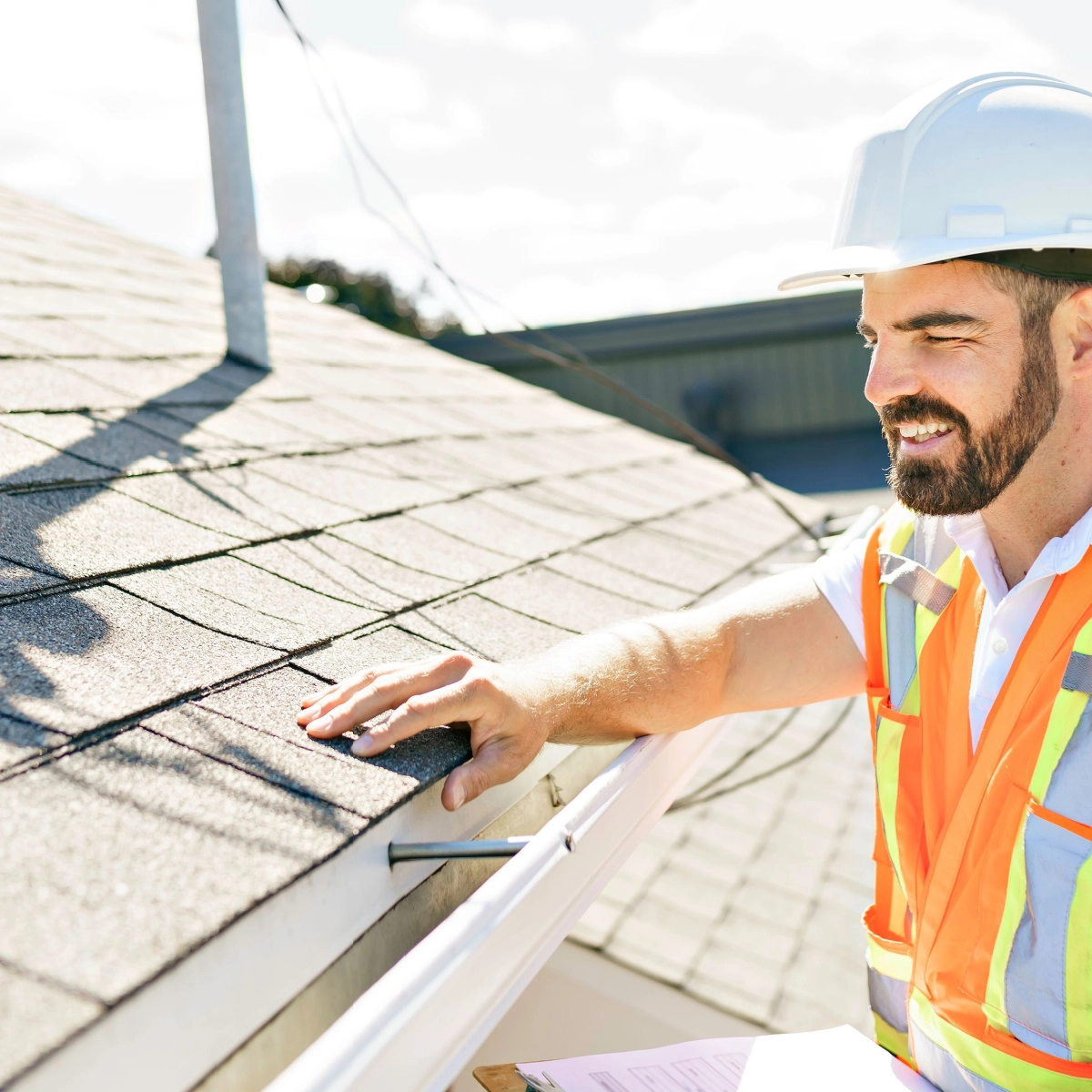 A man in a white hard hat and orange safety vest inspects a residential roof, presumably for a Tomball roof replacement. He has one hand on the shingles and a clipboard in the other. The background shows a clear sky and part of a building.