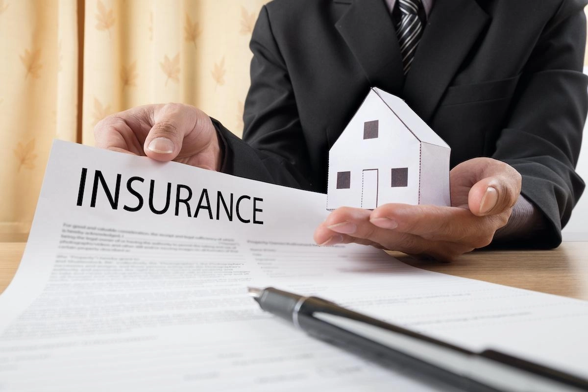 A person in a suit holds a small model of a house in one hand while pointing to an insurance document on a table, possibly discussing Houston roof replacement options. A pen rests on the document, emphasizing the concept of home insurance. The background features a blurred curtain.