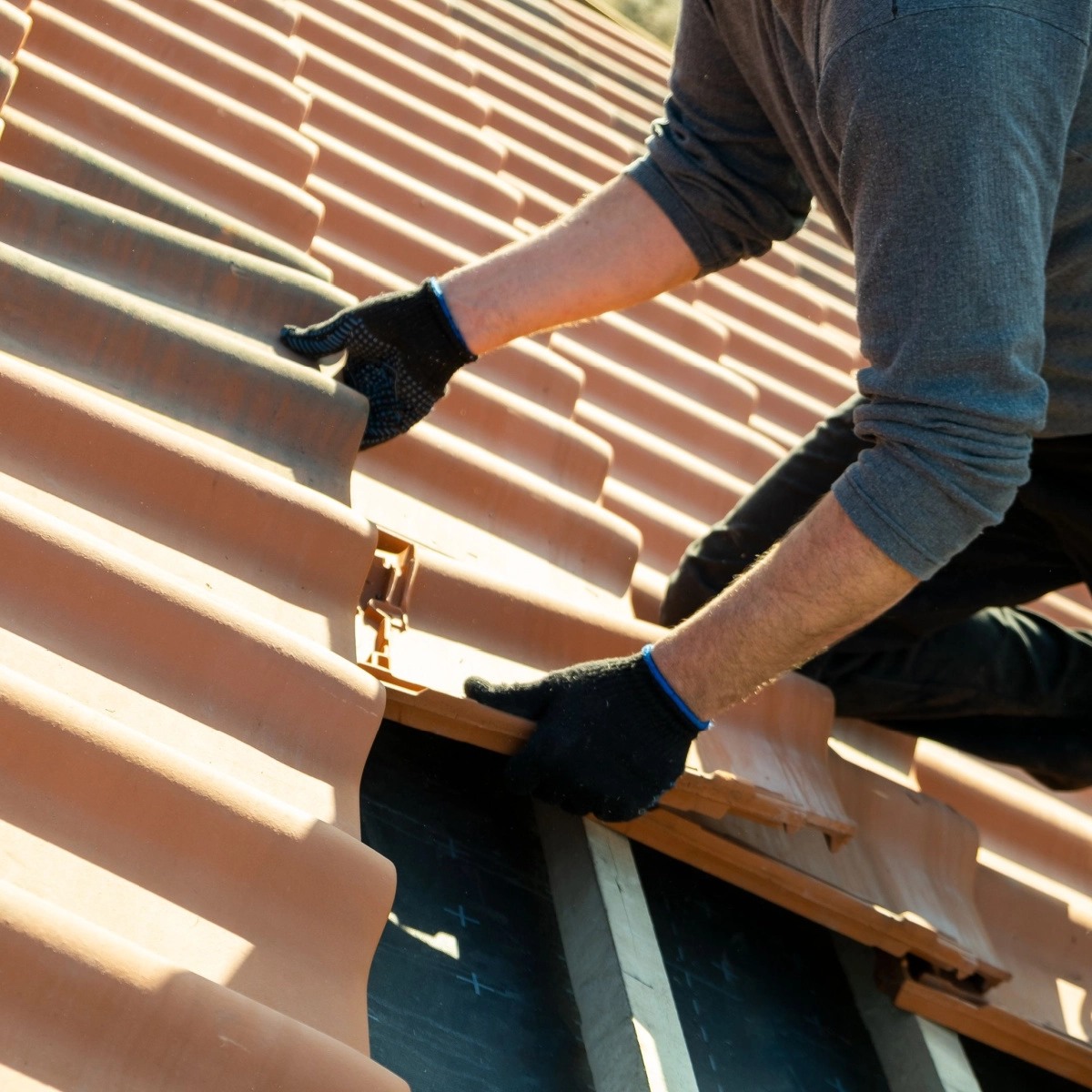 A person wearing a gray long-sleeve shirt and black gloves is installing or repairing red clay roof tiles in The Woodlands. They are kneeling on the roof, carefully positioning a tile. The person appears to be focused on aligning the tiles properly during the roof replacement.