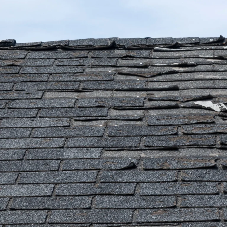 A close-up of an old, damaged shingle roof with multiple cracked, curled, and missing shingles, showing signs of significant wear and tear. The clear blue sky in the background highlights the urgent need for a Sugar Land roofer.