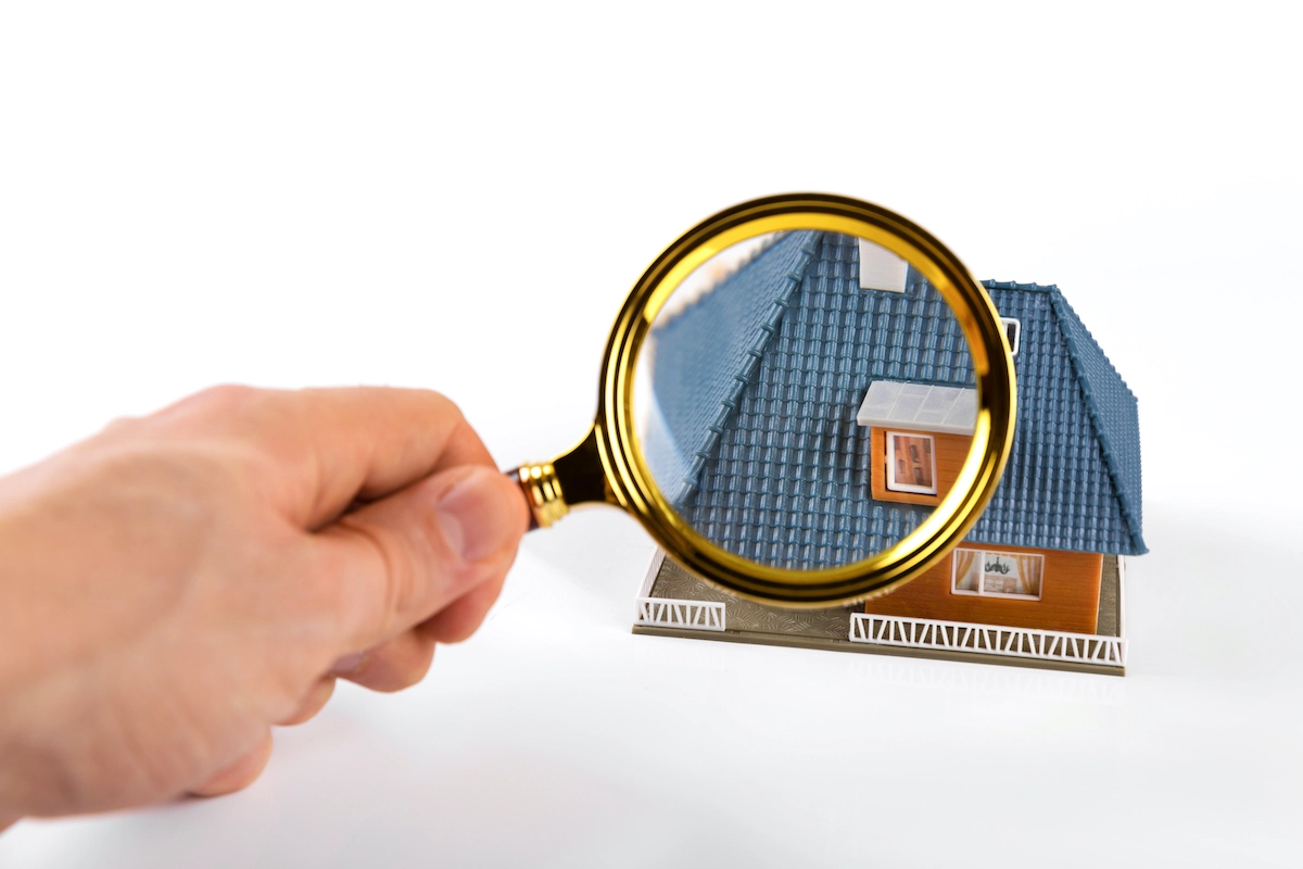 A hand holds a magnifying glass over a small model house with a blue roof, wooden walls, and a white fence around the perimeter. The magnifying glass zooms in on the details of the roof and part of a window, reminiscent of expert inspections by Houston roofing professionals. The background is plain white.