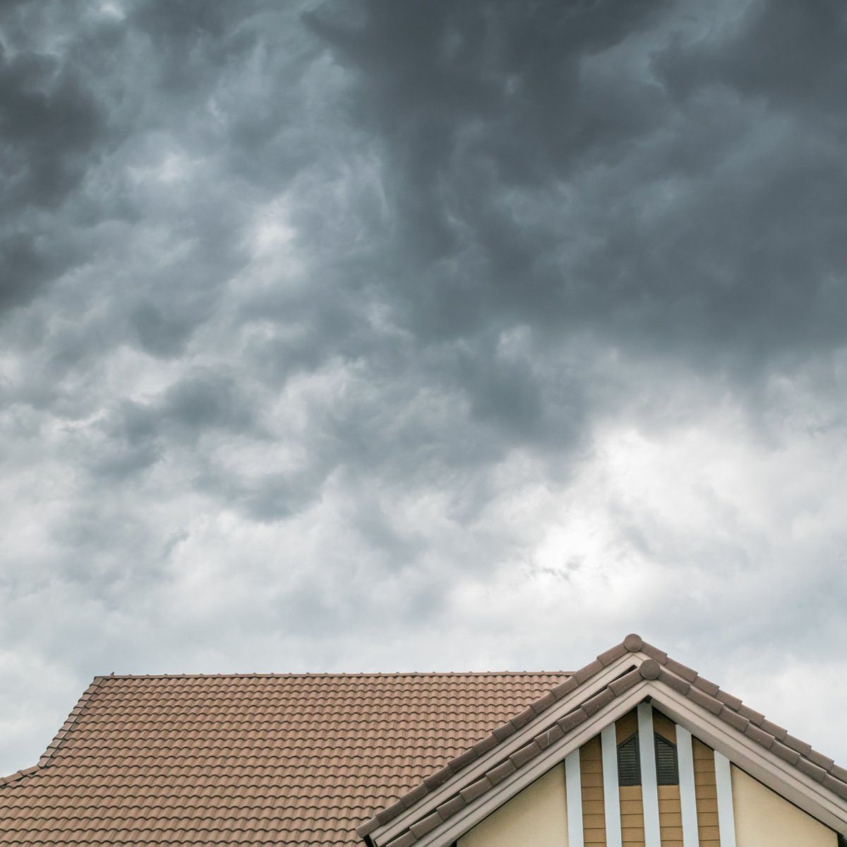 A house with a beige Friendswood roof and attic windows is shown under a cloudy, overcast sky with dark gray clouds suggesting an impending storm.