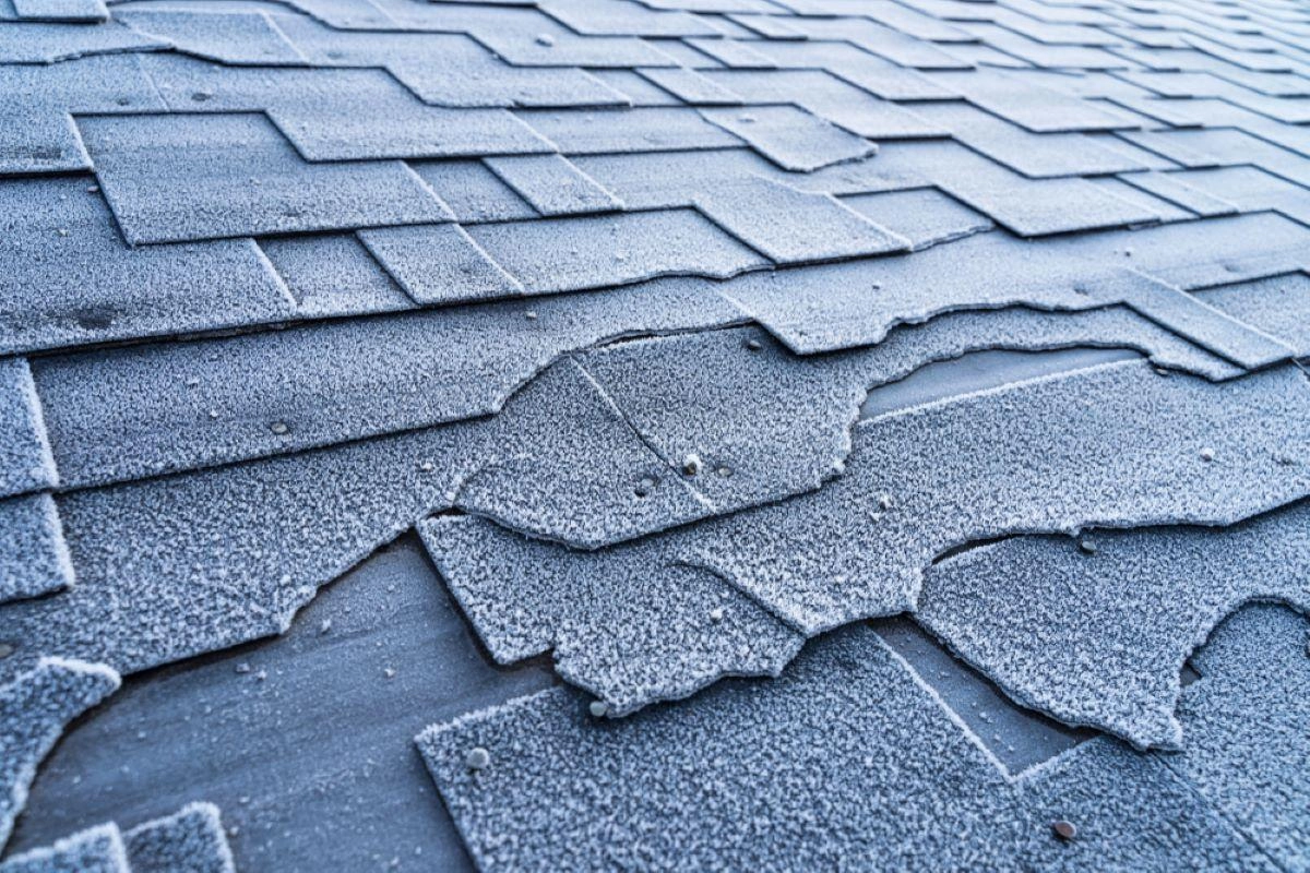 A close-up view of weathered, damaged roof shingles with irregular shapes and curled edges. The shingles appear frosty, indicating cold weather conditions. Some shingles are broken or missing, revealing the roof structure underneath—a typical concern for Spring TX roofing experts.