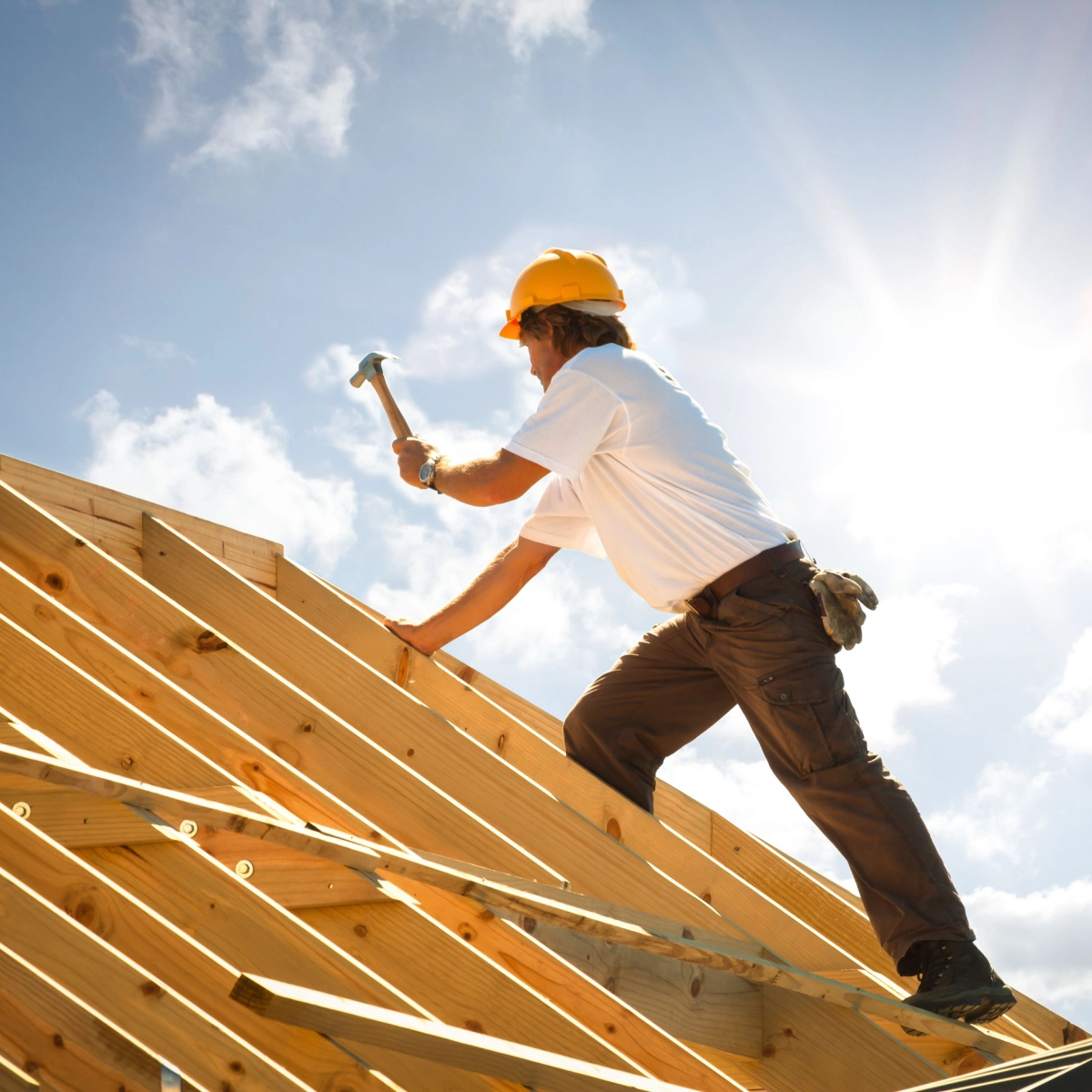 A construction worker in a yellow hard hat and white shirt hammers nails into a wooden roof frame under a bright, sunny sky with scattered clouds. The worker, part of the Bellaire roof replacement team, stands on the wooden beams with the sun shining brightly in the background.