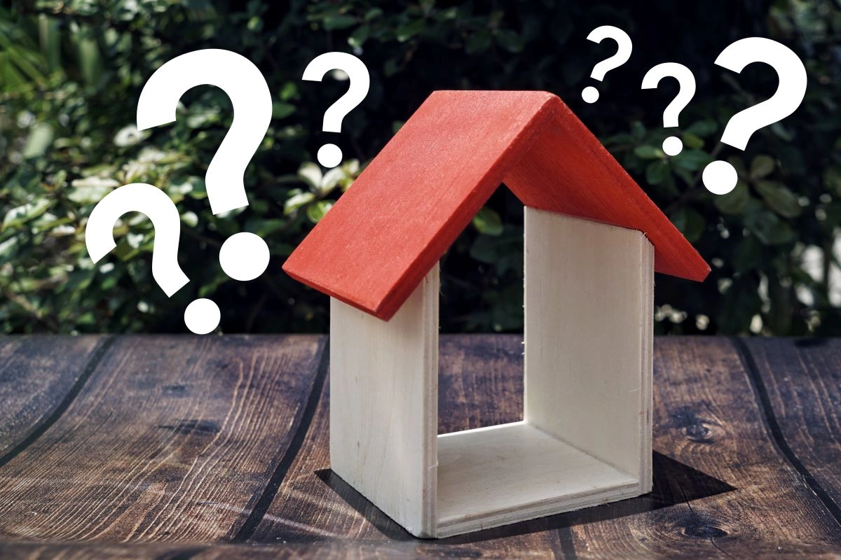 A small wooden model of a house with a red roof stands on a dark wooden table. Large white question marks are superimposed around the house against a leafy green background, suggesting confusion or uncertainty relating to home or real estate concepts, possibly hinting at The Woodlands roof replacement issues.