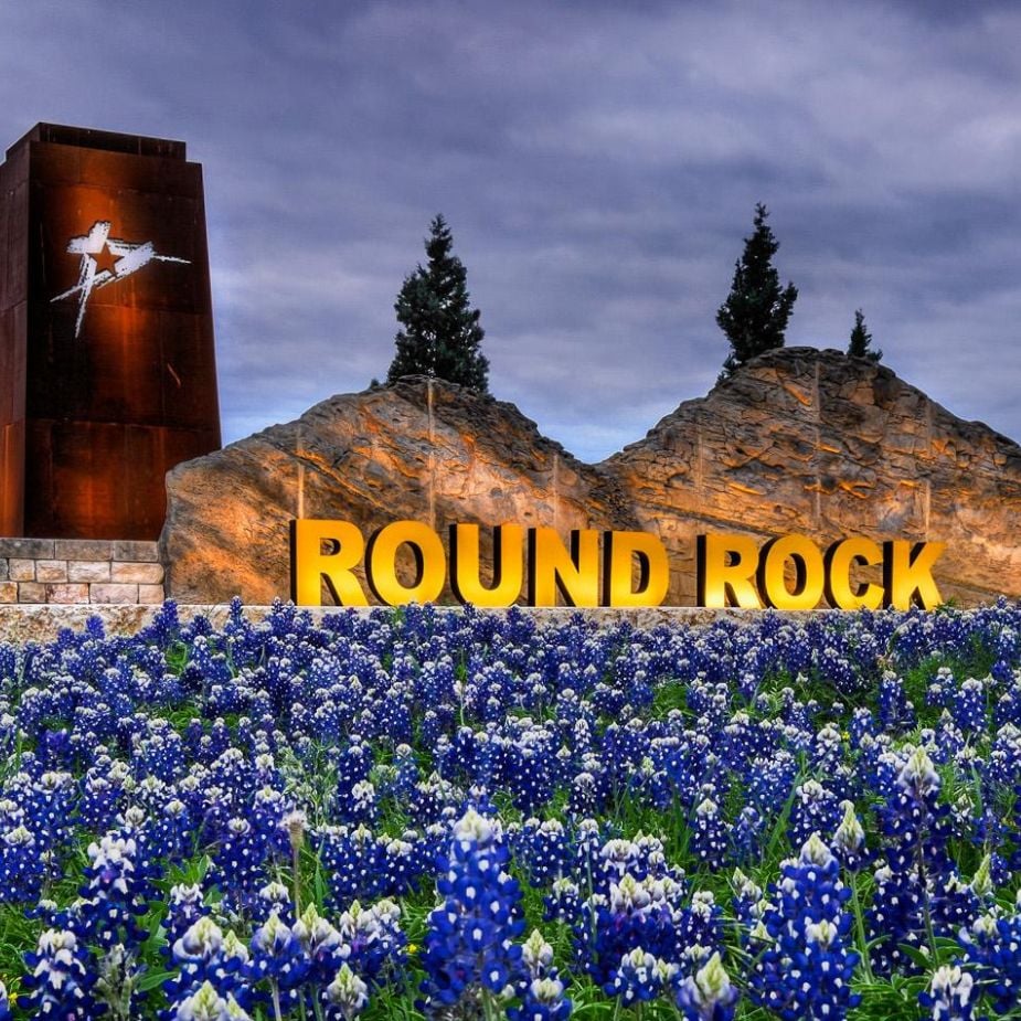 A large sign reading "ROUND ROCK" is set against a backdrop of rocky terrain and tall trees. The sign, glowing brightly, stands prominently above a field of vibrant bluebonnets under a cloudy sky, symbolizing the resilience and craftsmanship you'd expect from a top roofing contractor in Round Rock.