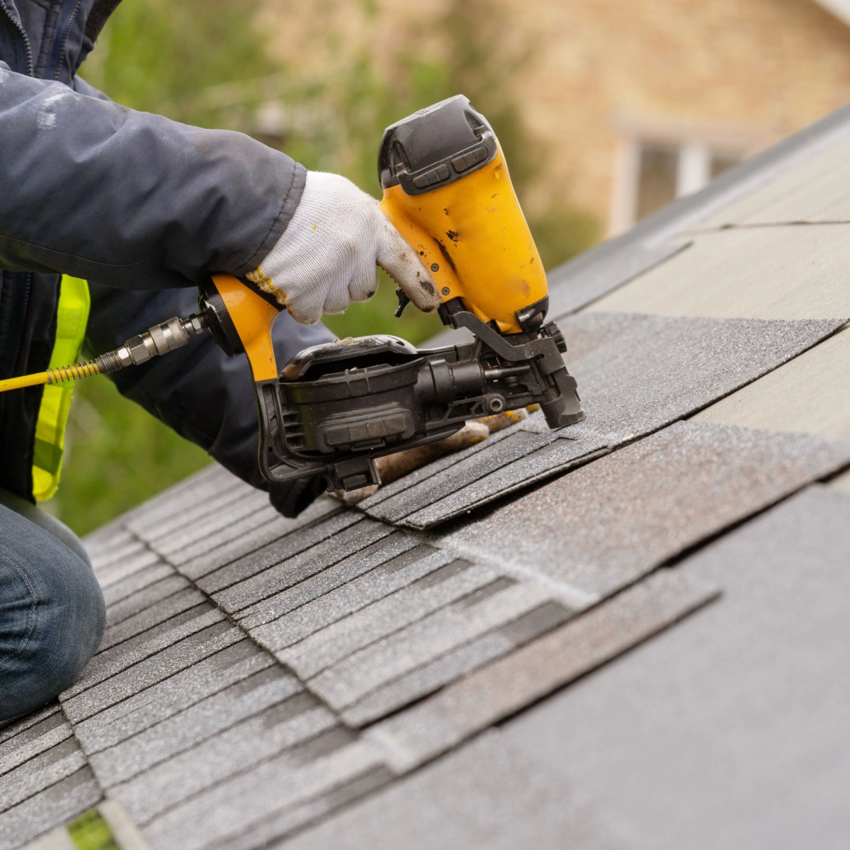 A person in work gloves uses a nail gun to secure asphalt shingles on a roof. The individual, dressed in a jacket, is kneeling on the sloped surface, carefully aligning the shingles. In the background, blurred greenery and part of a building hint at The Woodlands roof replacement project.