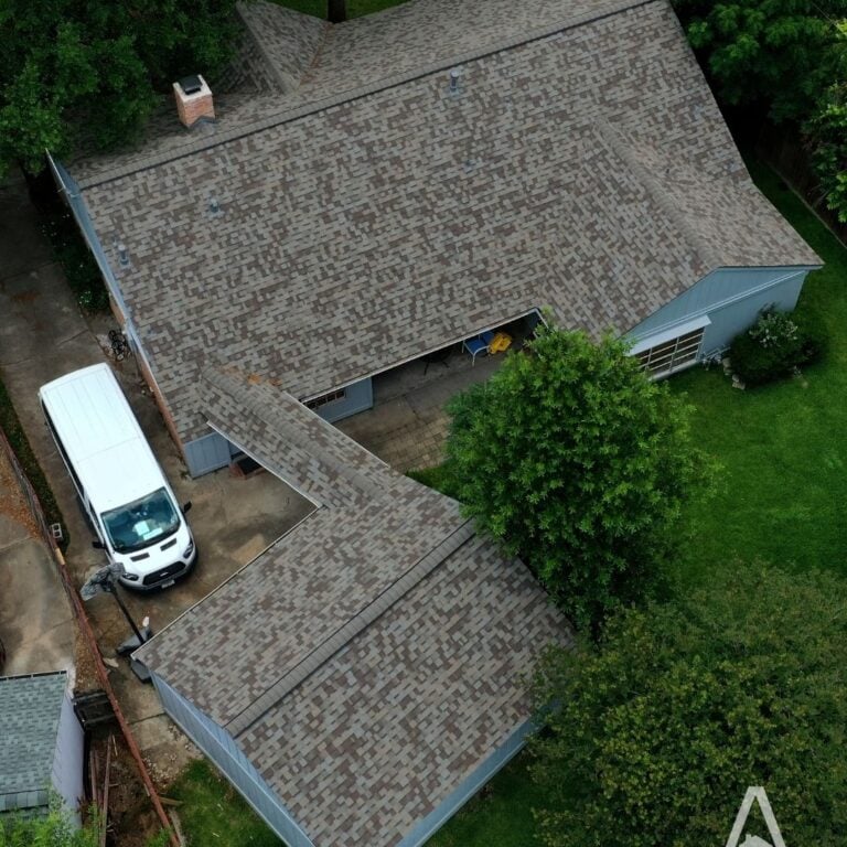 Aerial view of a house with a gray shingled rooftop, expertly done by a roofing contractor in Houston, surrounded by green trees. A white van is parked in the driveway adjacent to the house. The property features a large backyard with patches of vibrant grass.