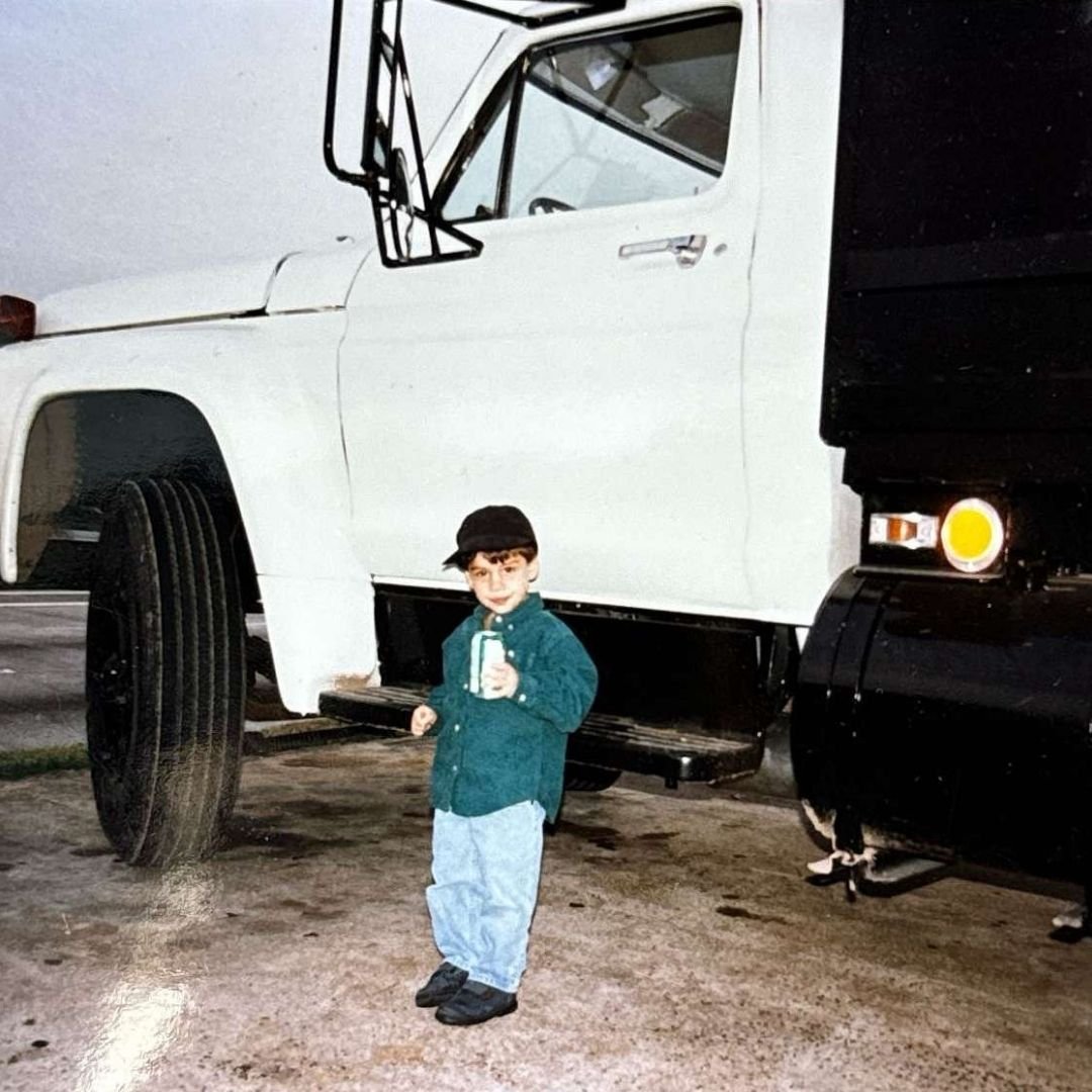 A young child wearing a dark cap, green shirt, and jeans stands beside a large white truck with a black trailer from a roofing contractor in Houston. The child is holding a can and smiling at the camera. The background appears to be an outdoor parking area.
