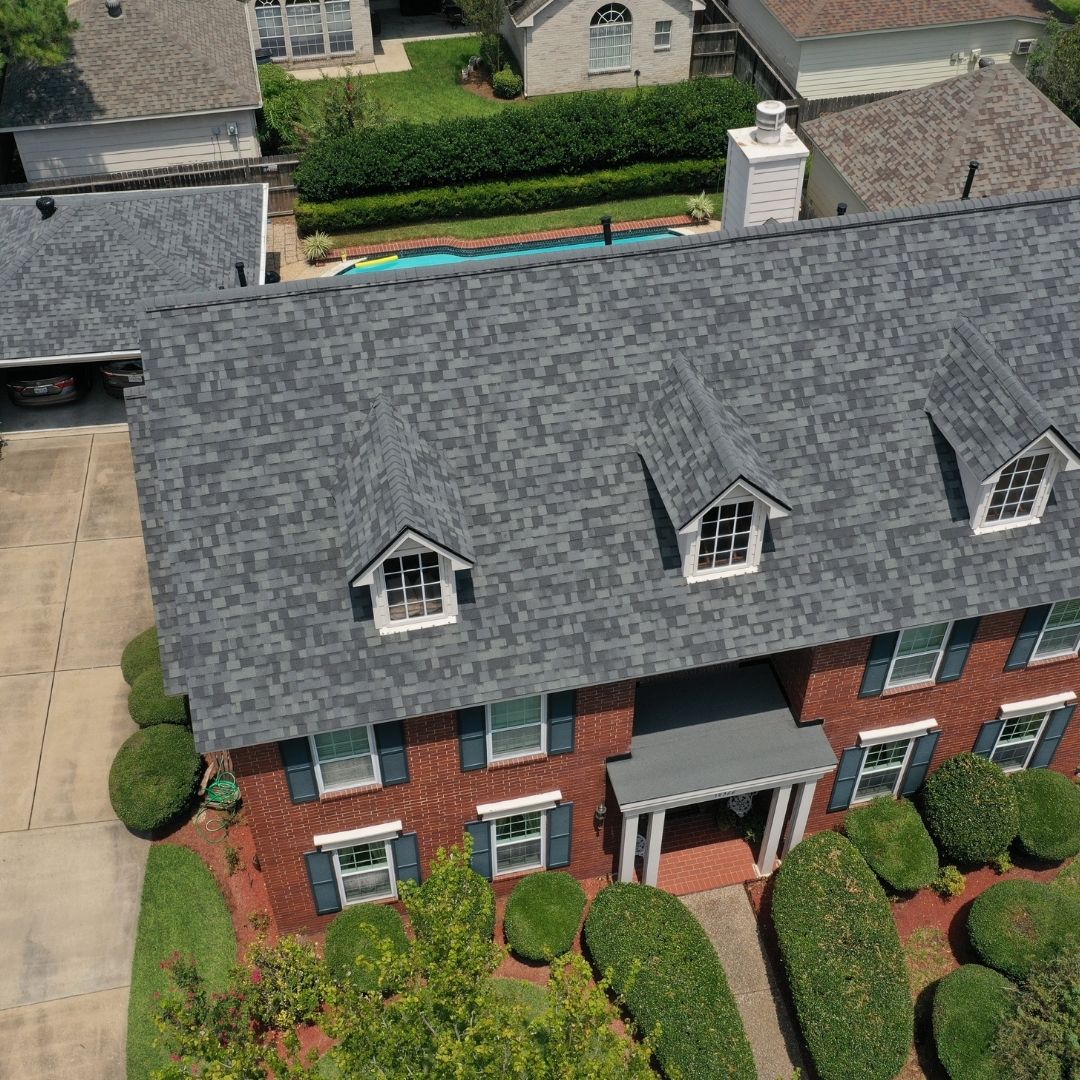 Aerial view of a two-story red-brick house with a grey shingle roof, crafted by a top roofing company in Memorial City. It features dormer windows, neatly trimmed hedges, and a paved driveway. A backyard pool peeks into view amidst nearby houses and greenery.