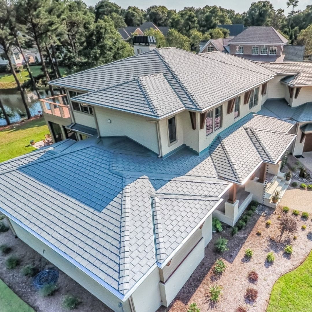 Aerial view of a large, modern house with an intricate, multi-tiered roof structure expertly crafted by a roofing company in Memorial City. The property is surrounded by a well-maintained garden with landscaped pathways. Nearby, there is a water body reflecting the greenery of the surrounding trees.