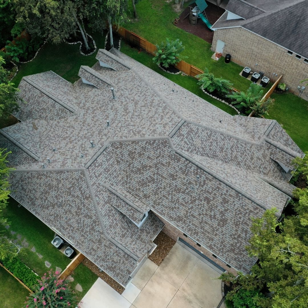Aerial view of a suburban two-story house with a complex, multi-gabled roof design. The house is surrounded by trees, a manicured lawn, and has a wooden fence in the backyard. Another house is visible in the right-side background. The driveway is seen at the bottom—perfect work from a roofing company in Memorial City.