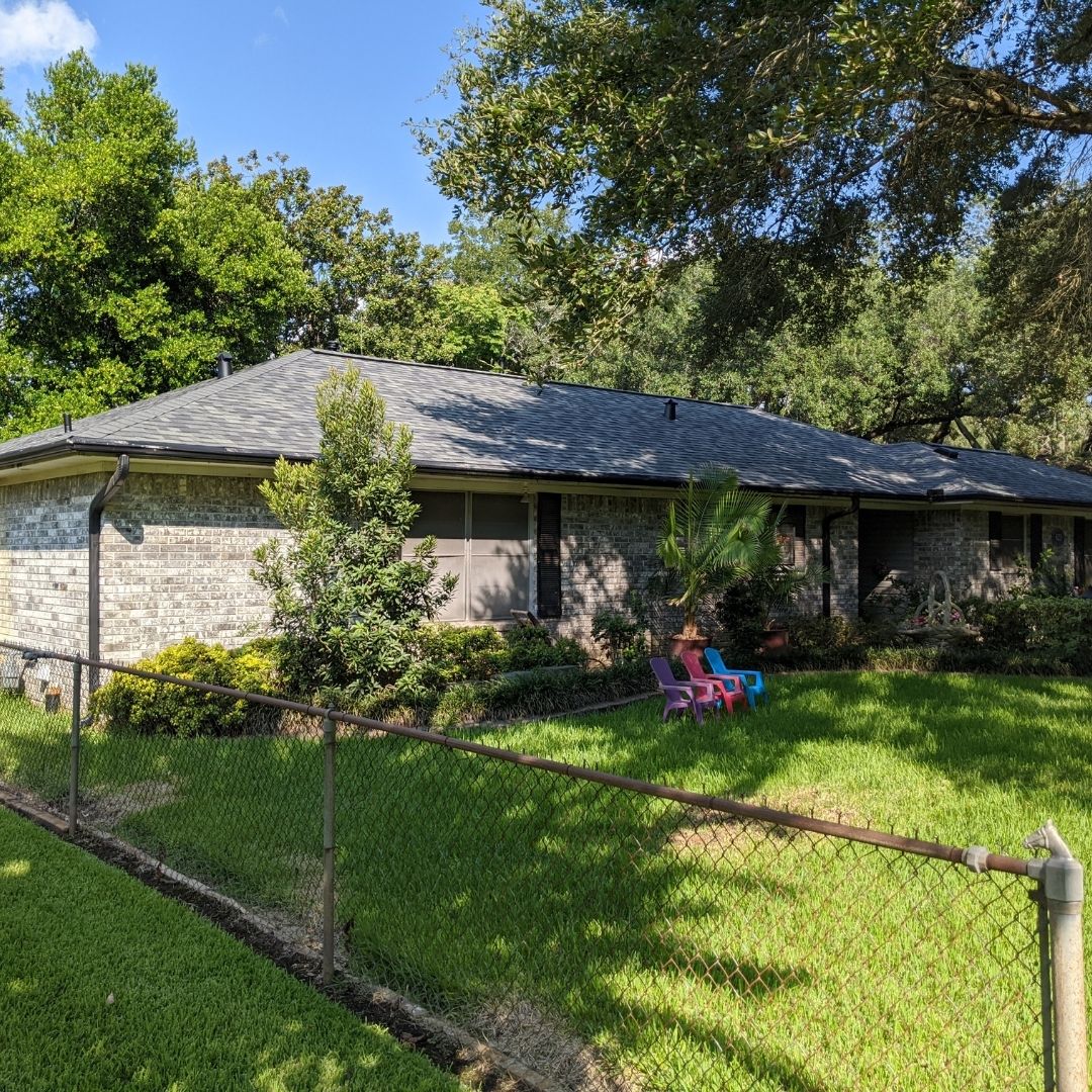 A single-story brick house with a grey roof, likely installed by a top roofing company in Memorial City, is surrounded by well-maintained greenery. The front yard features a chain-link fence, several trees, shrubs, and colorful outdoor chairs on the lawn. Bright and sunny weather enhances the lush, green landscape.