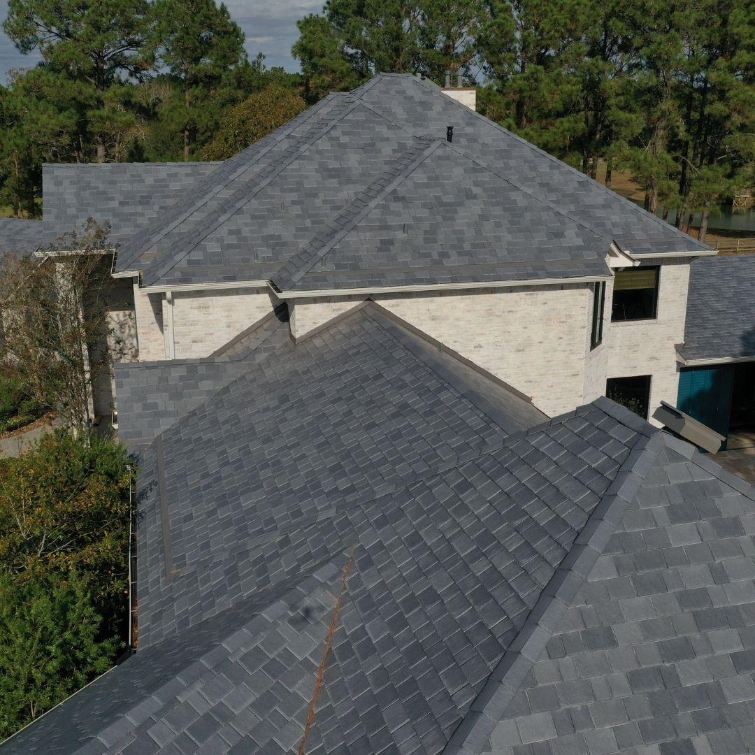 A large, multi-level house with light brick walls is shown. The home's roofing, expertly crafted by a top roofing company in Memorial City, is covered with grey asphalt shingles arranged in a detailed pattern, highlighting the complex and varied angles of the roof. Surrounding the house are tall trees and a clear sky above.