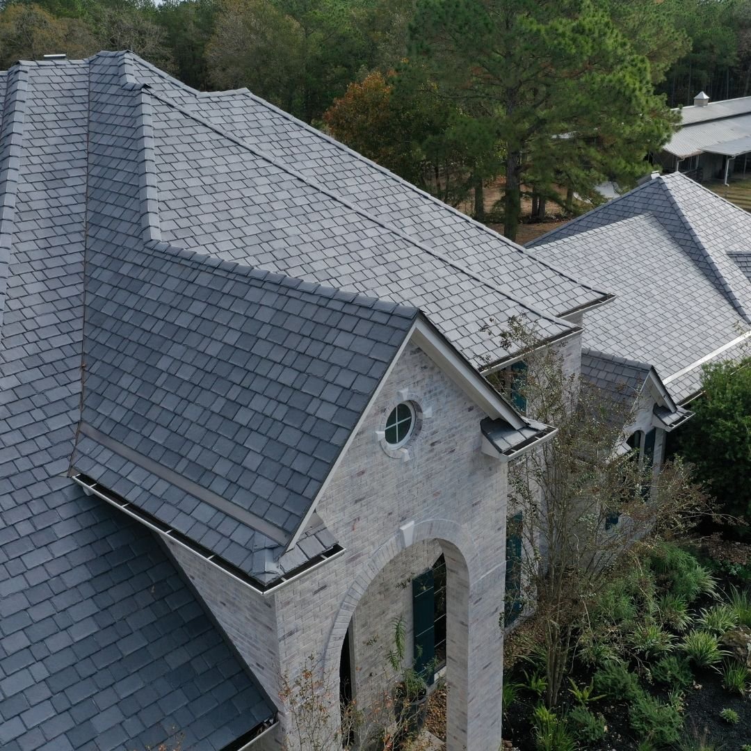 Aerial view of a large house with a gray shingled roof, crafted by a renowned roofing company in Memorial City. The structure features multiple gables and arched windows, surrounded by trees and greenery.