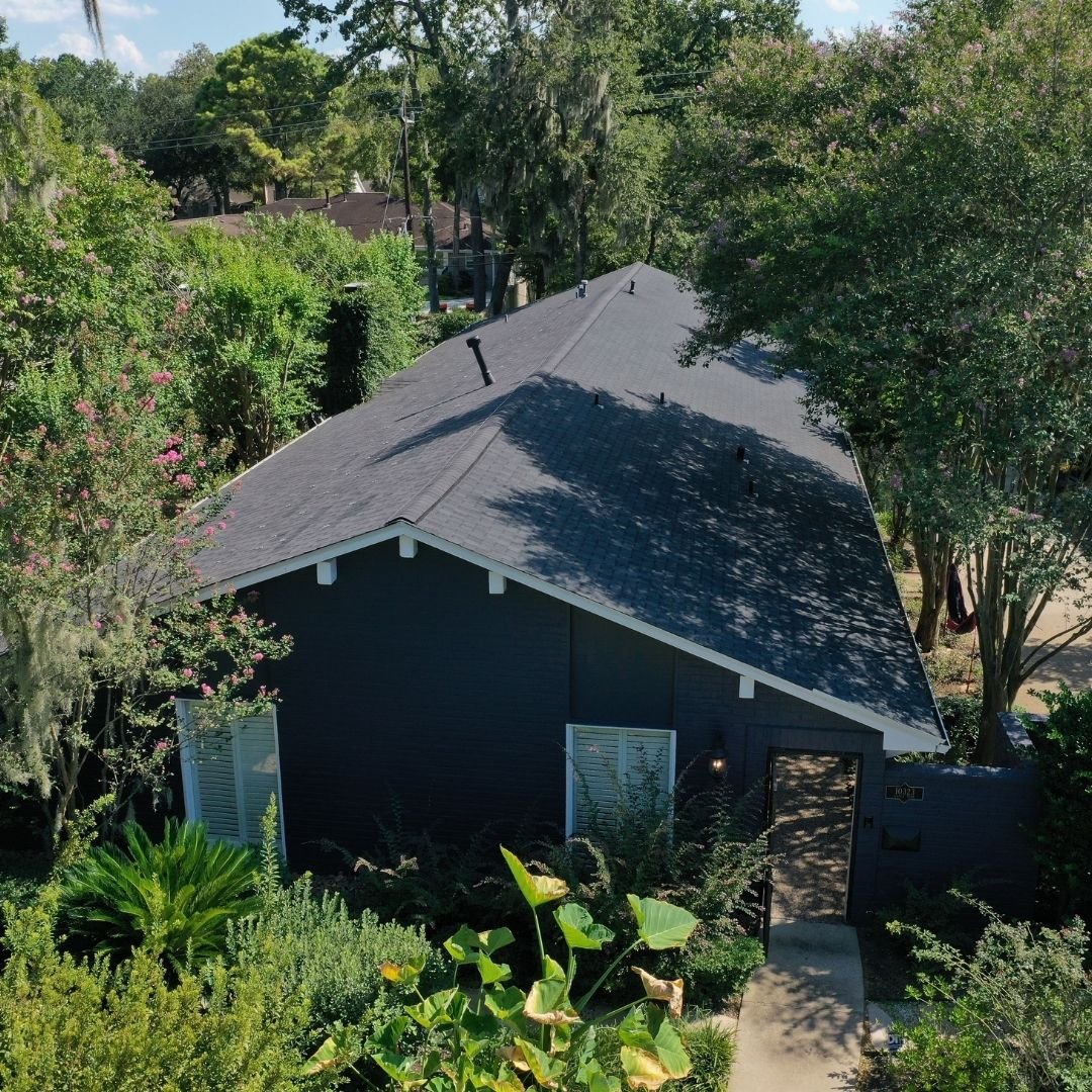Aerial view of a dark gray house with a steep gabled roof, expertly crafted by a roofing company in Memorial City, partially obscured by lush green trees and plants. The house has white trim and sits in a densely vegetated area, creating a secluded and serene atmosphere.